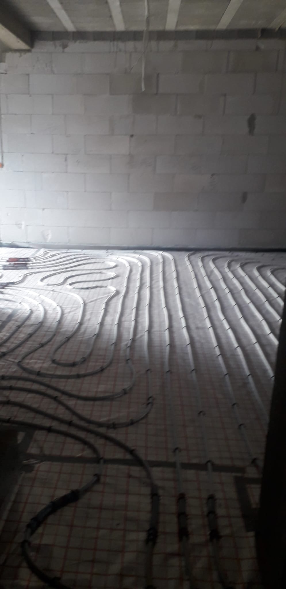 Ułożone rury ogrzewania podłogowego na siatce w surowym wnętrzu. Widoczne białe ściany z betonu komórkowego i szare rury tworzące pętle na podłodze.