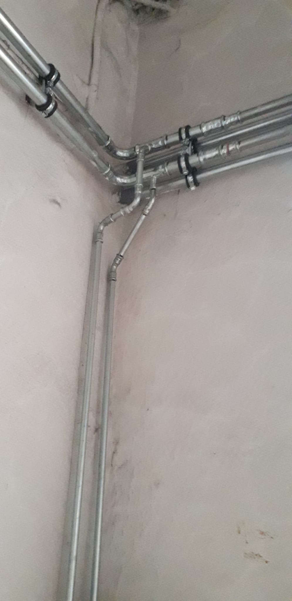 Szare, metalowe rury instalacji, przymocowane do ściany z widocznymi śladami wilgoci w rogu pomieszczenia. Rury prowadzone pod różnymi kątami, z licznymi złączkami i mocowaniami.