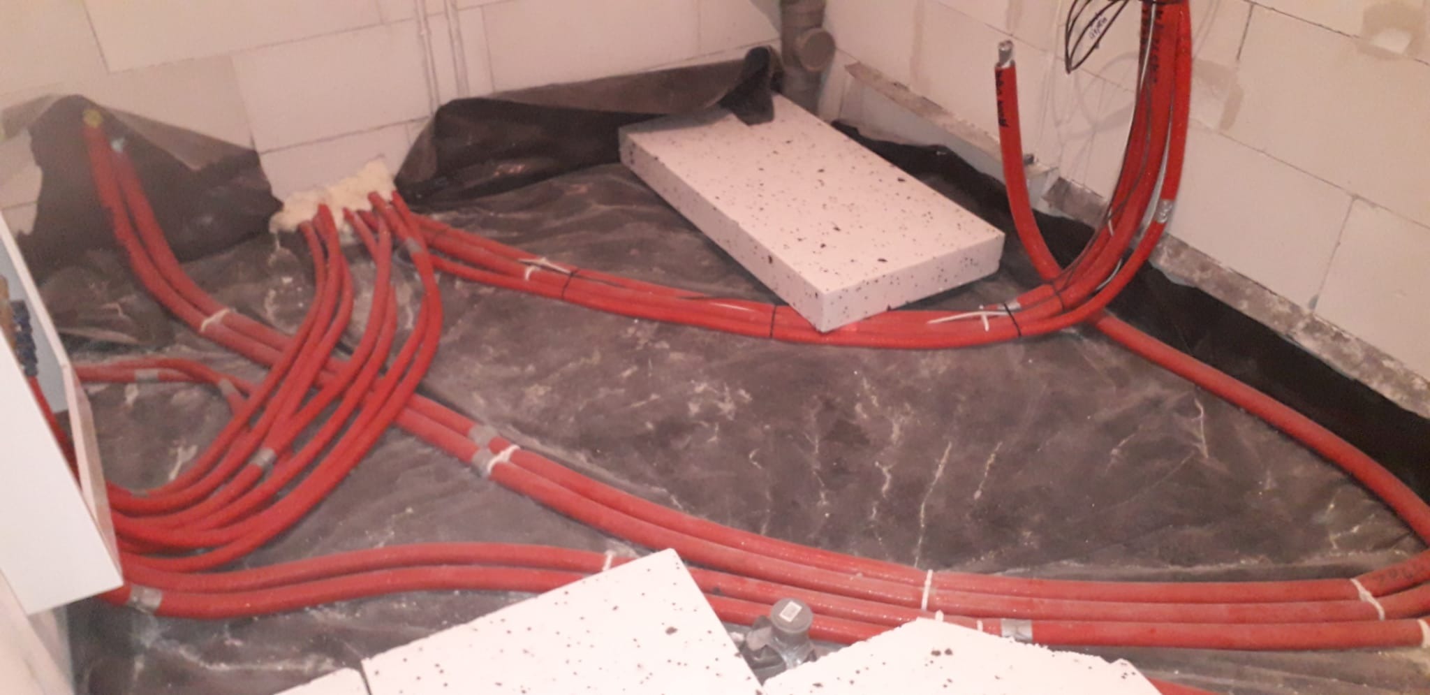 Czerwone rury ogrzewania podłogowego, częściowo przymocowane do podłoża, z widocznymi białymi płytami izolacyjnymi i folią ochronną na podłożu.