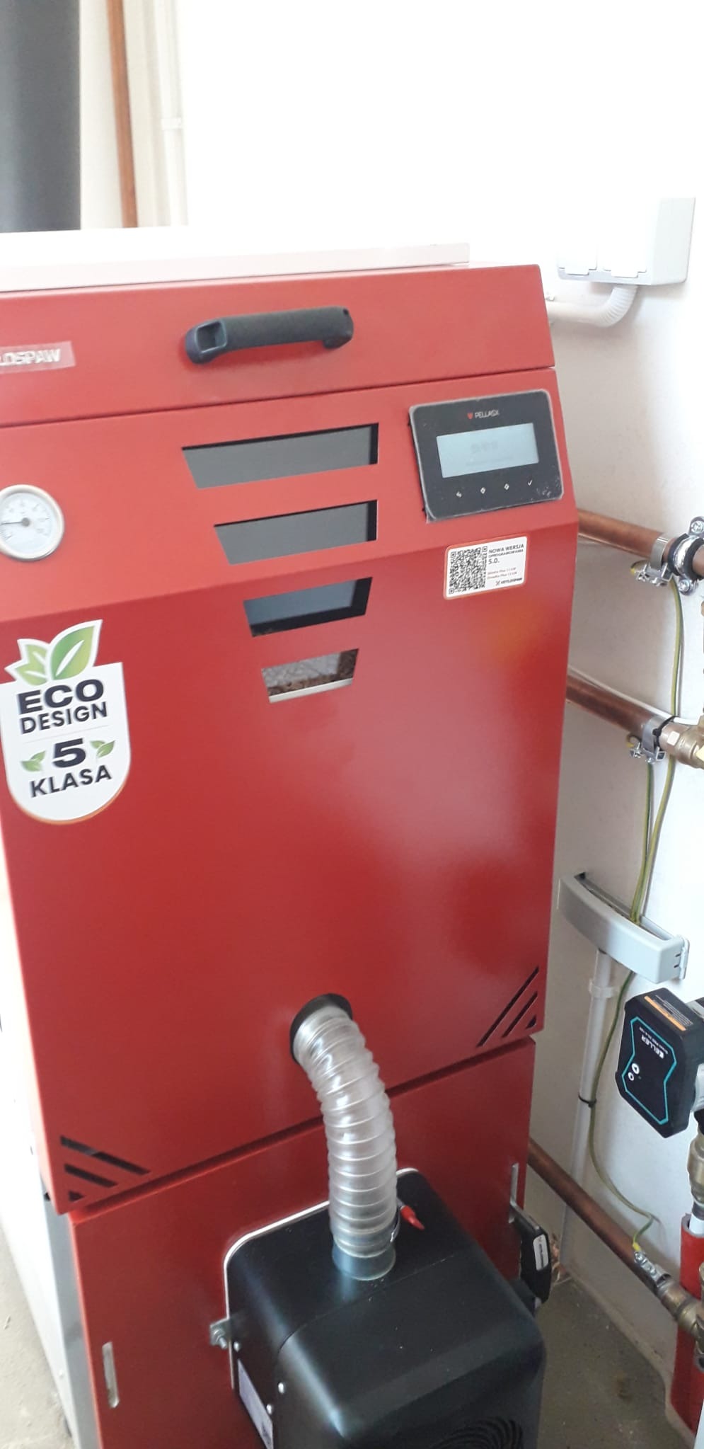 Czerwony piec na pellet z wyświetlaczem, naklejką Eco Design 5 Klasa i podłączonym przewodem wentylacyjnym. Widoczne rury instalacji grzewczej.