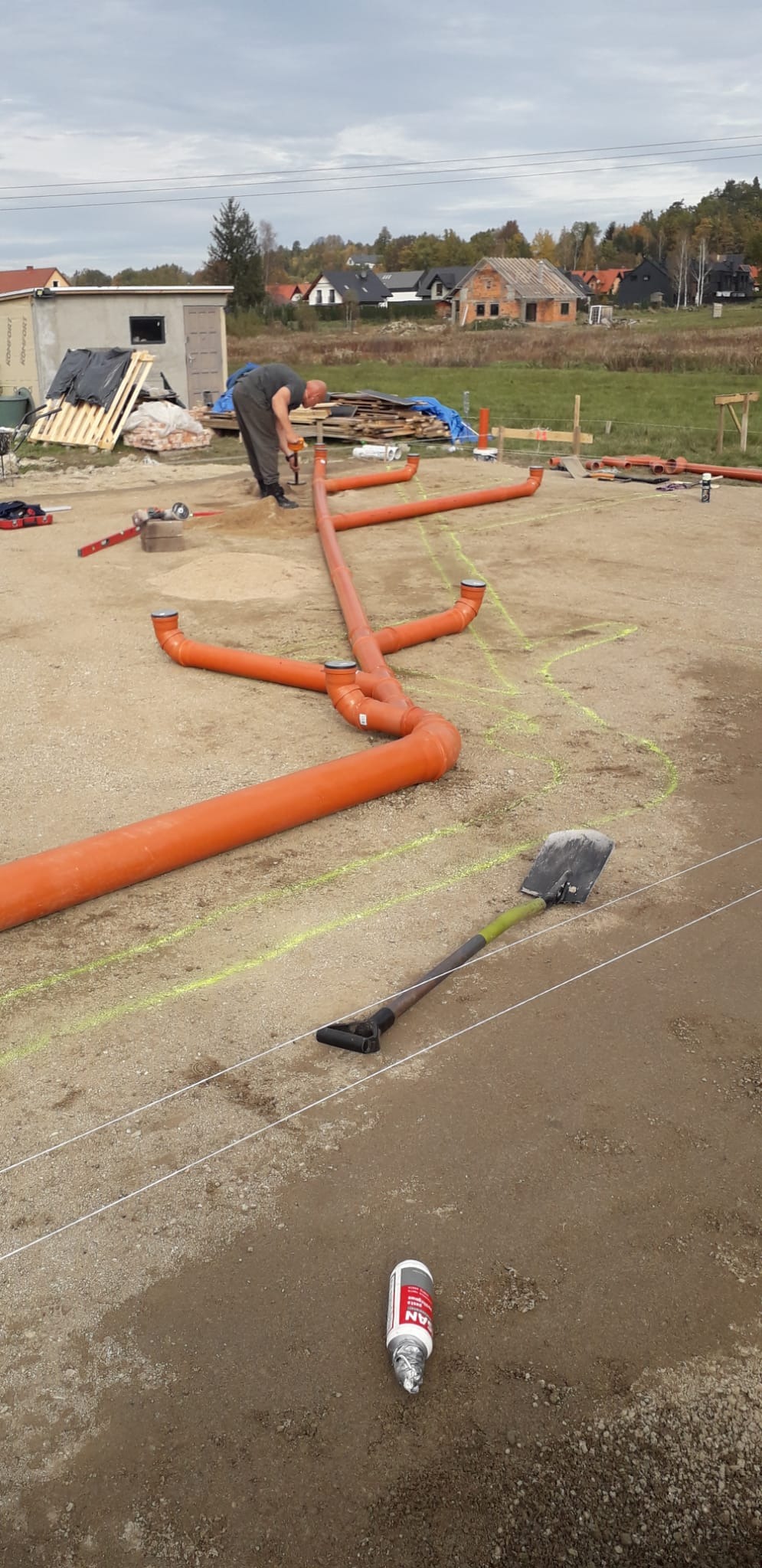 Montaż pomarańczowych rur kanalizacyjnych na piasku, widoczny pracownik i narzędzia. Rury ułożone w ziemi, przygotowane do dalszych prac. Tło stanowią domy w oddali.