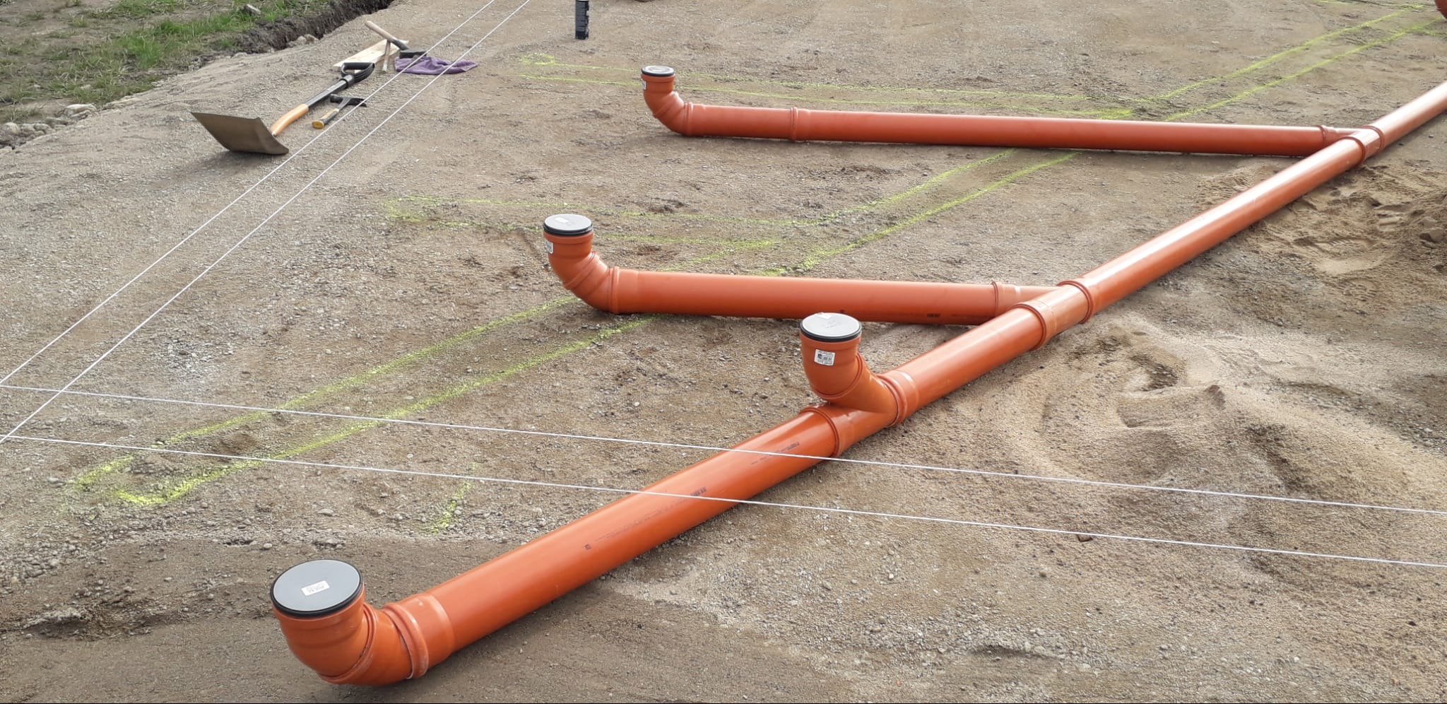 Pomarańczowe rury kanalizacyjne z odpowietrznikami ułożone na piasku, wytyczone sznurkiem. W tle widoczna łopata i inne narzędzia.