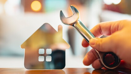Metalowy model domu i klucz nastawny w dłoni na drewnianym blacie. Symbol napraw, konserwacji nieruchomości, zarządzania budynkiem. Rozmyte tło.