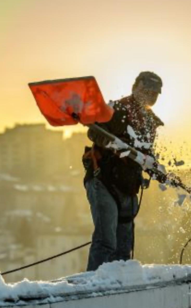 Pracownik w uprzęży usuwa śnieg z dachu budynku za pomocą czerwonej łopaty. W tle widoczne budynki i zachodzące słońce.