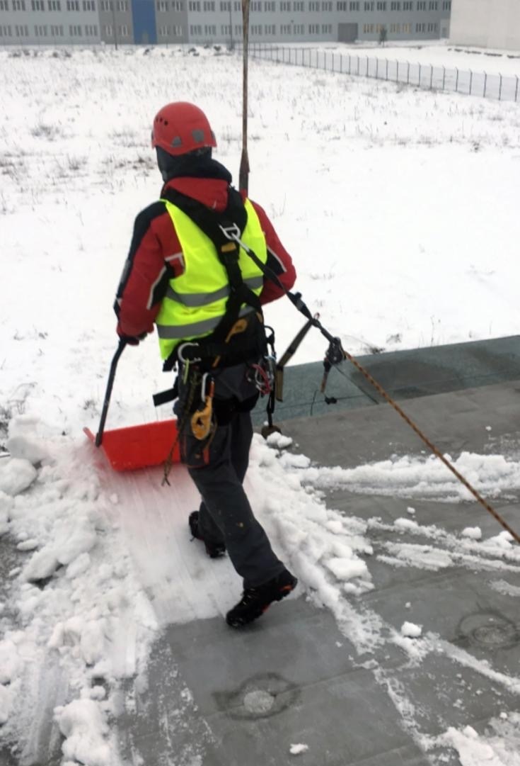 Pracownik w kasku i uprzęży odśnieża płaski dach budynku przemysłowego za pomocą czerwonej łopaty. W tle ośnieżone pole i budynek.