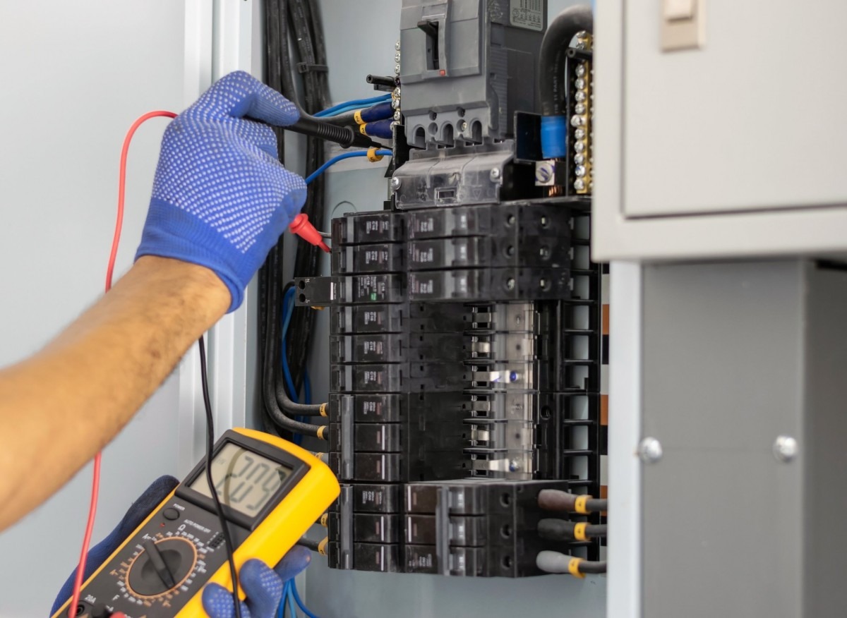 Sprawdzanie napięcia w skrzynce elektrycznej przez elektryka w niebieskich rękawicach, używającego multimetru z żółtą obudową. Widoczne kable i bezpieczniki.