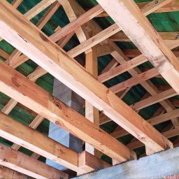 Konstrukcje drewniane Kamil Hewig - Widok od dołu na drewnianą konstrukcję więźby dachowej z zieloną membraną, betonowy komin w tle.
