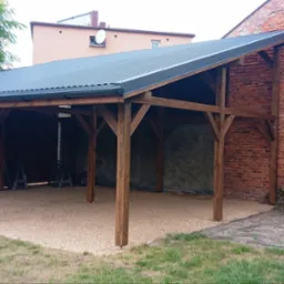 Drewniana konstrukcja wiaty samochodowej z ciemnym, metalowym dachem, wsparta na solidnych, brązowych słupach, z podłożem wysypanym drobnym żwirem, przylegająca do ceglanego budynku i otoczona...
