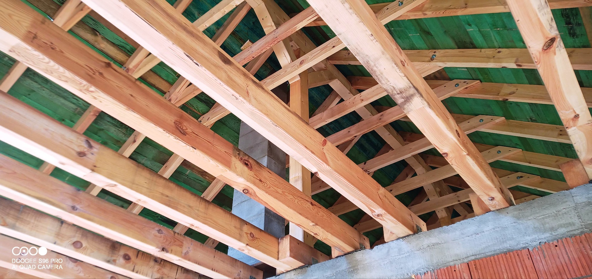 Widok od dołu na drewnianą konstrukcję więźby dachowej z zieloną membraną, betonowy komin w tle.