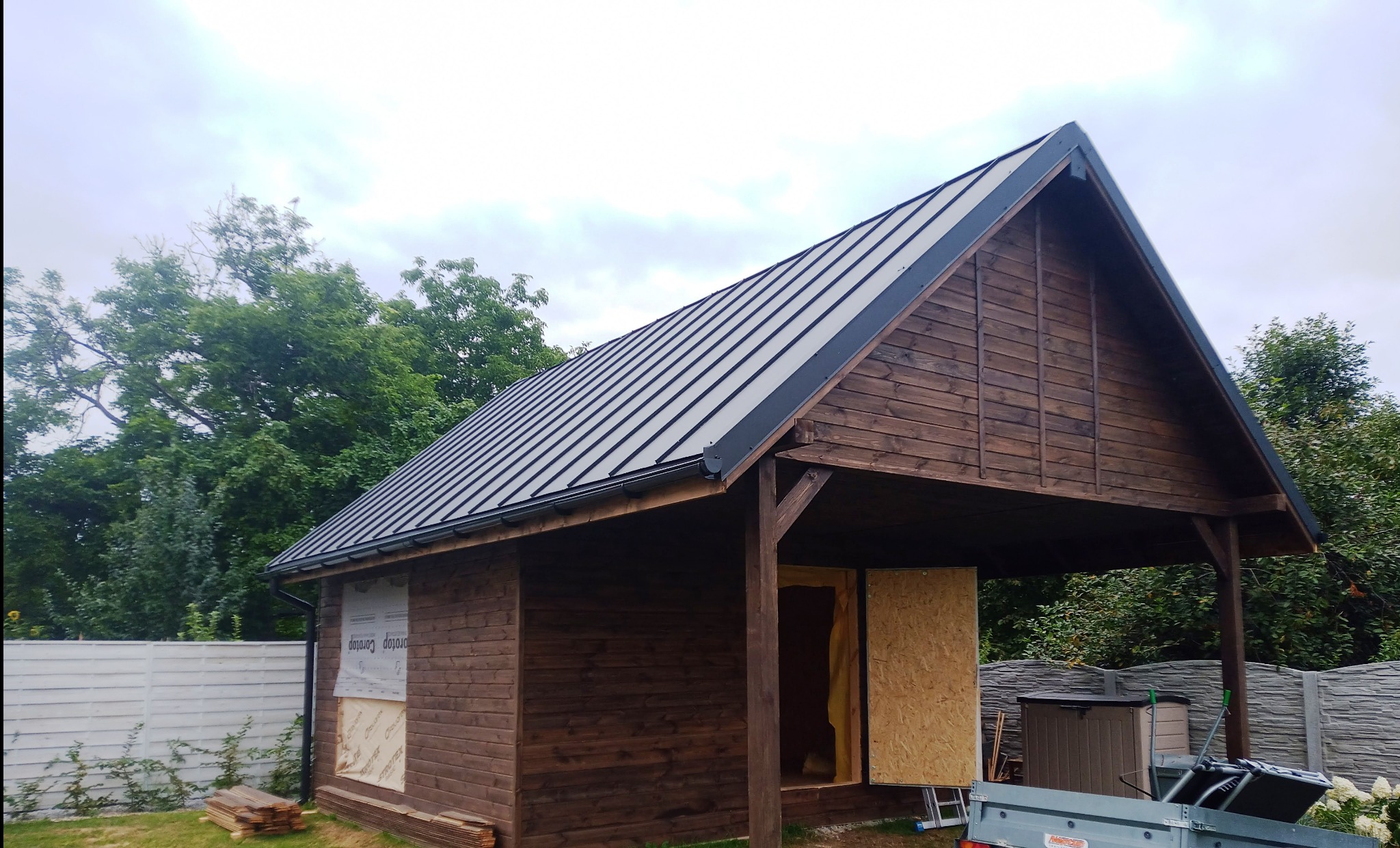 Drewniana konstrukcja z czarnym, metalowym dachem w trakcie budowy, widoczne deski i tymczasowe zabezpieczenia okien, z przyczepą i narzędziami w tle, na tle zieleni.