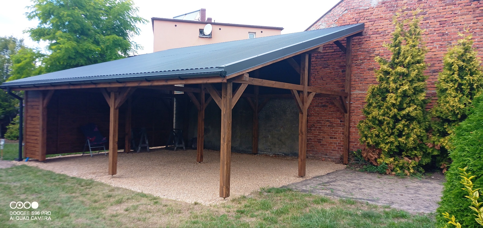 Drewniana konstrukcja wiaty samochodowej z ciemnym, metalowym dachem, wsparta na solidnych, brązowych słupach, z podłożem wysypanym drobnym żwirem, przylegająca do ceglanego budynku i otoczona...