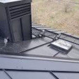 Centrum Dachowe Żyrardów S.C. - Szary dach z blachy trapezowej, widoczny komin z czarną obudową, okno dachowe i elementy systemu odwadniającego. Dach pokryty warstwą wilgoci. Tło stanowią drzewa bezlistne.