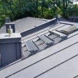 Centrum Dachowe Żyrardów S.C. - Szary dach z blachy trapezowej z kominem i oknem dachowym, widok z góry na tle zieleni drzew.