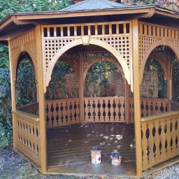 Fachurex - Drewniana altana ogrodowa po malowaniu, widoczne dwie puszki farby na podłodze, świeżo pomalowane elementy konstrukcyjne, tło z zieleni.