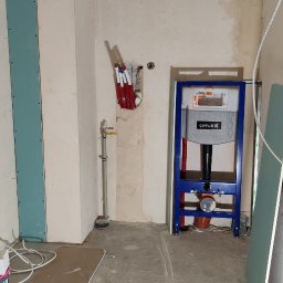 Fachurex - Łazienka w trakcie remontu: widoczne płyty karton-gips na ścianach, stelaż podtynkowy WC marki Cersanit w kolorze niebieskim, rury instalacyjne wystające ze ściany, oraz częściowo ułożona podłoga.