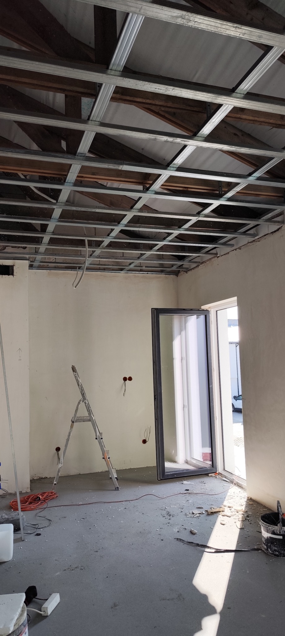 Wnętrze w trakcie prac wykończeniowych, widoczna konstrukcja sufitu podwieszanego z profili metalowych, drabina, otwór okienny i drzwiowy, ślady po pracach na podłodze.