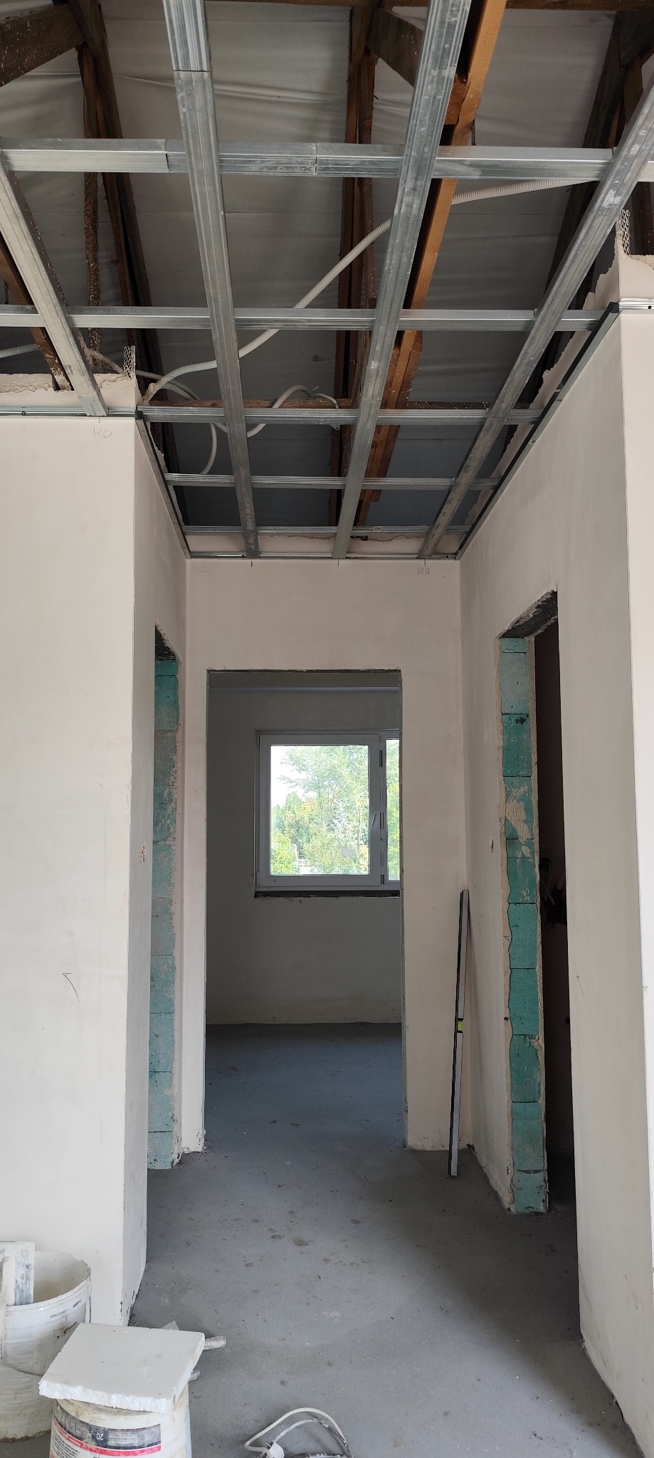Wnętrze w trakcie prac wykończeniowych: widoczne metalowe profile sufitowe przed montażem płyt kartonowo-gipsowych, ściany częściowo otynkowane, surowa podłoga, otwory drzwiowe w trakcie obróbki.