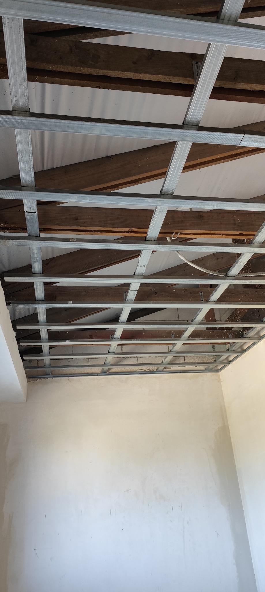 Metalowa konstrukcja sufitu podwieszanego z płyt kartonowo-gipsowych, widoczne drewniane belki stropowe i fragmenty instalacji elektrycznej, ściany w stanie surowym.