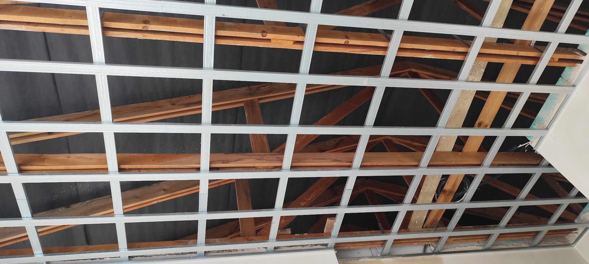 Metalowa konstrukcja sufitu podwieszanego z widocznymi drewnianymi elementami więźby dachowej i czarną folią izolacyjną w tle.