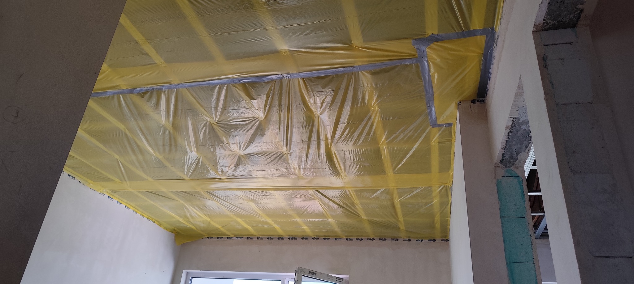 Montaż folii paroizolacyjnej na drewnianej konstrukcji sufitu podwieszanego, z użyciem taśmy uszczelniającej, widok z dołu na surowy stan pomieszczenia.