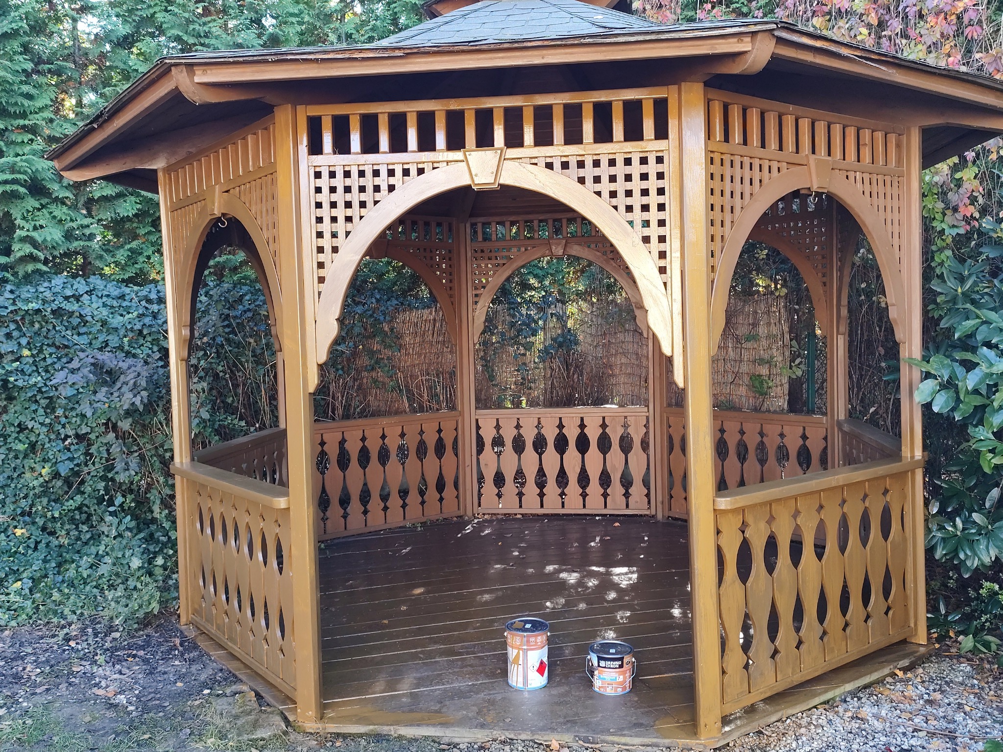Drewniana altana ogrodowa po malowaniu, widoczne dwie puszki farby na podłodze, świeżo pomalowane elementy konstrukcyjne, tło z zieleni.