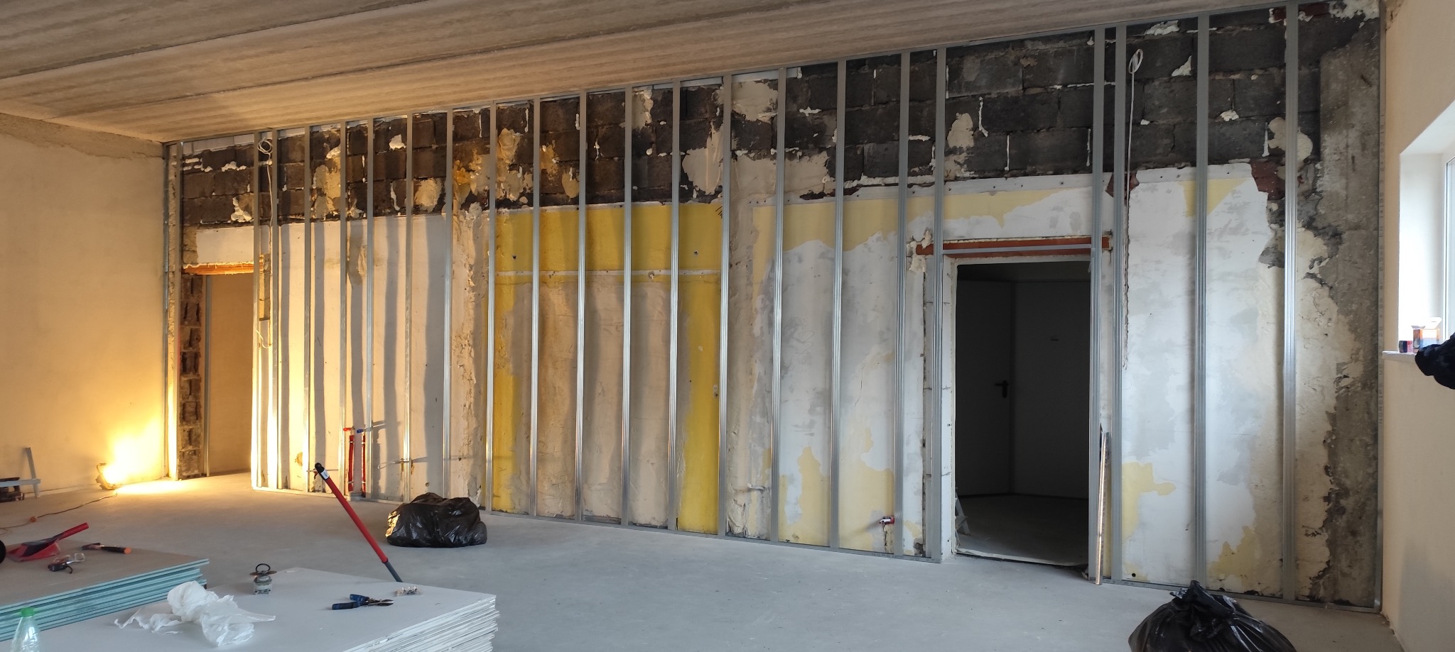 Montaż metalowego rusztu pod płyty gipsowo-kartonowe na ścianie z widocznymi fragmentami starej żółtej tapety i instalacji, z otworami drzwiowymi po obu stronach, na podłodze leżą płyty...