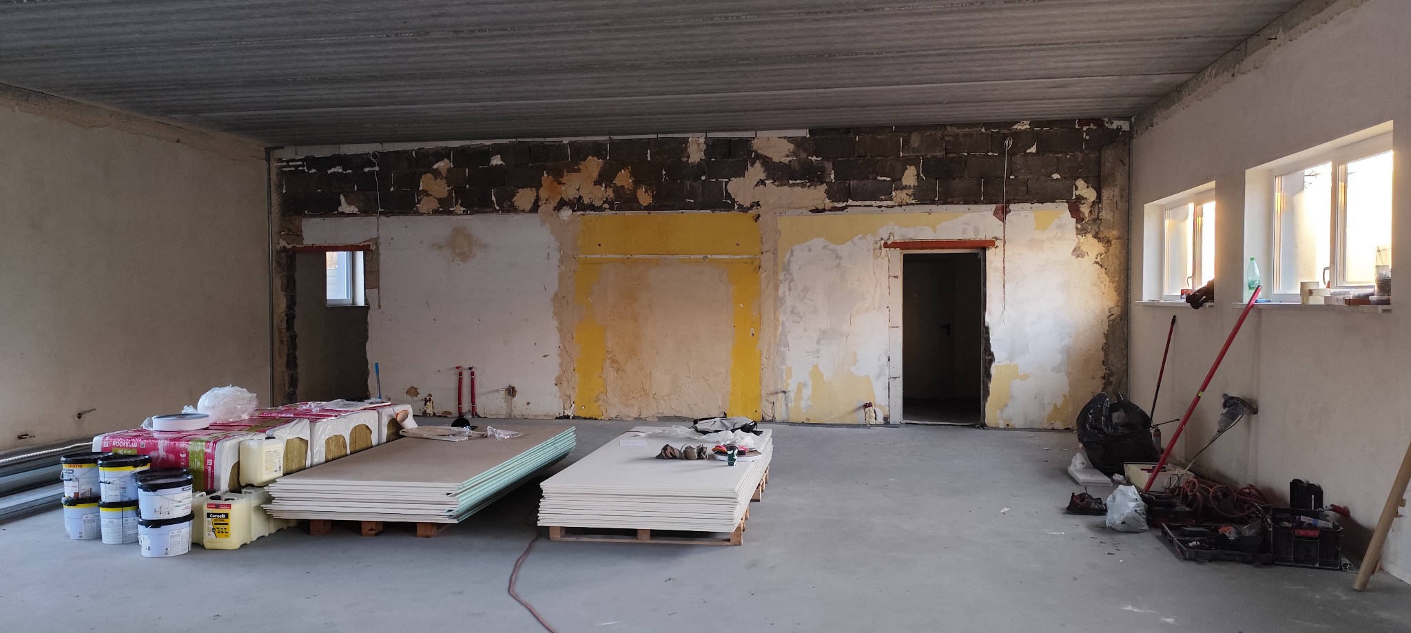 Wnętrze w trakcie prac: widoczne płyty karton-gips na paletach, materiały izolacyjne, puszki z farbą, odsłonięte fragmenty ściany z resztkami farby, dwa okna z widocznym światłem dziennym.