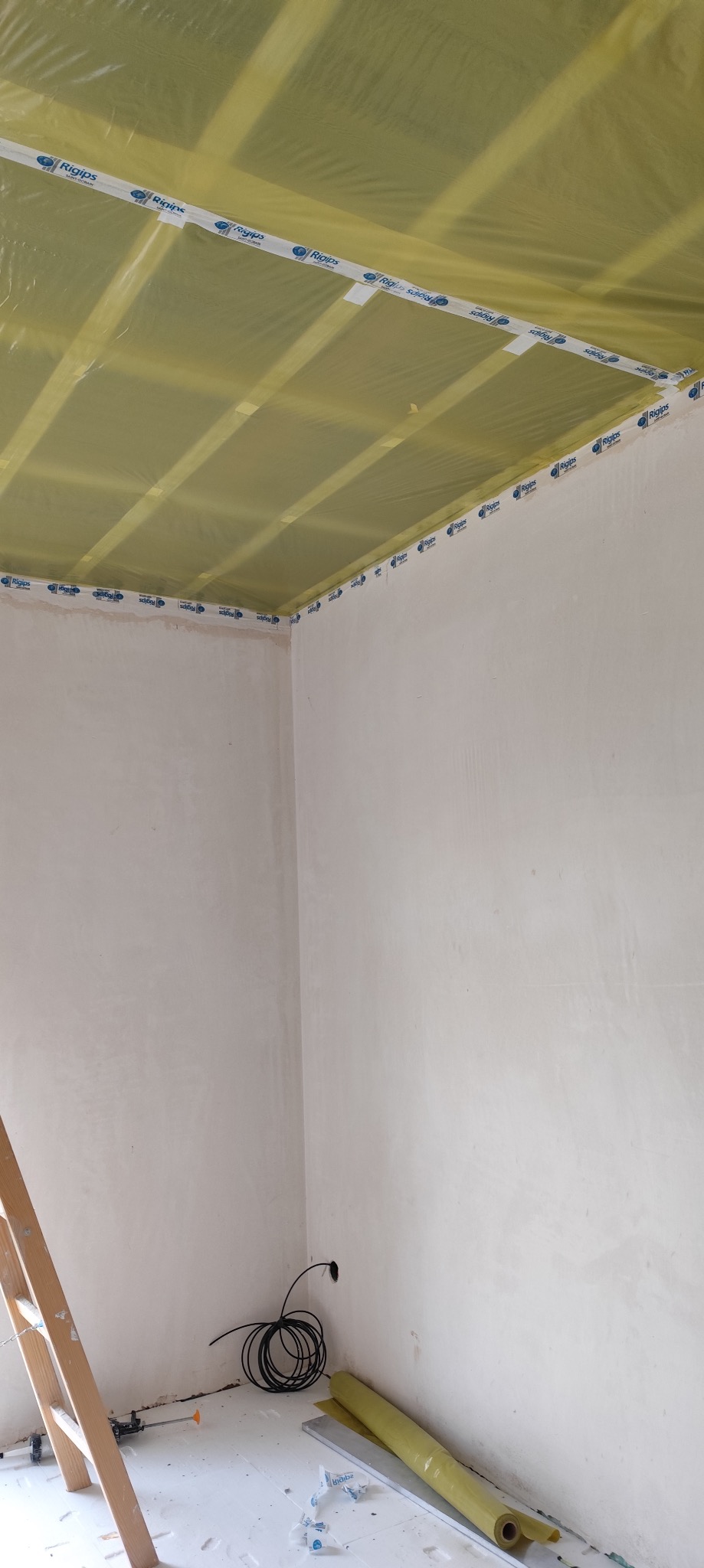 Przygotowanie do montażu sufitu podwieszanego: widoczne profile Rigips zabezpieczone folią, taśma uszczelniająca na obrzeżach ścian, przewody elektryczne wystające ze ściany, drabina oparta o ścianę.