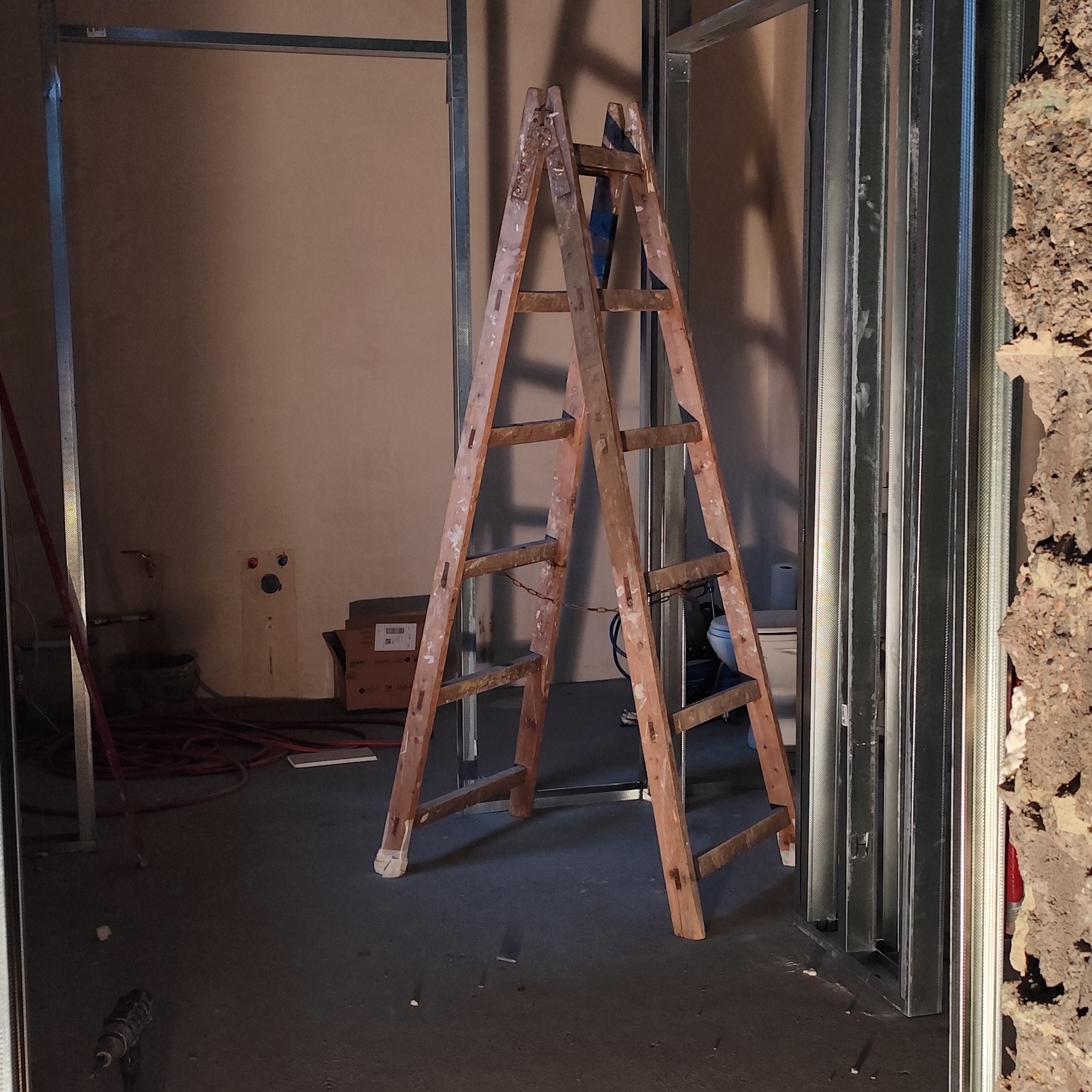Wnętrze w trakcie prac wykończeniowych: drewniana drabina oparta o metalowe profile konstrukcyjne, widoczne instalacje hydrauliczne, karton z etykietami w tle, fragment ściany z odsłoniętym murem...