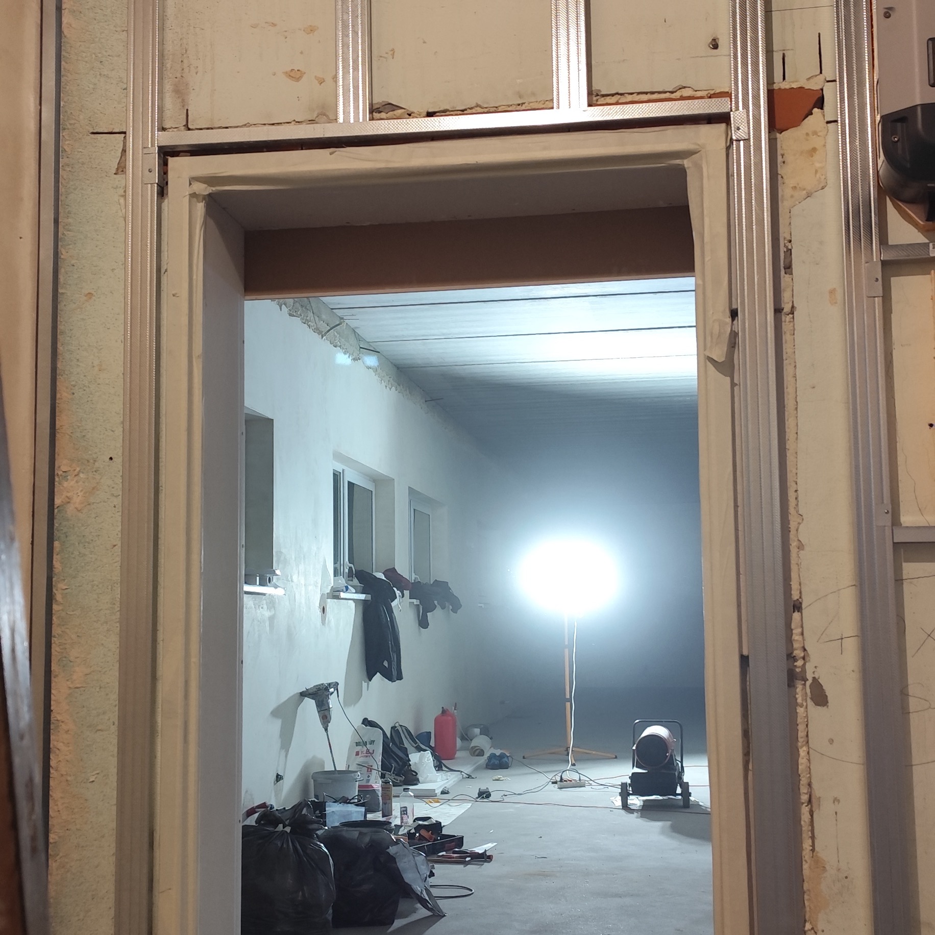 Montaż ościeżnicy w surowym wnętrzu z widocznymi profilami metalowymi, narzędziami i lampą budowlaną.