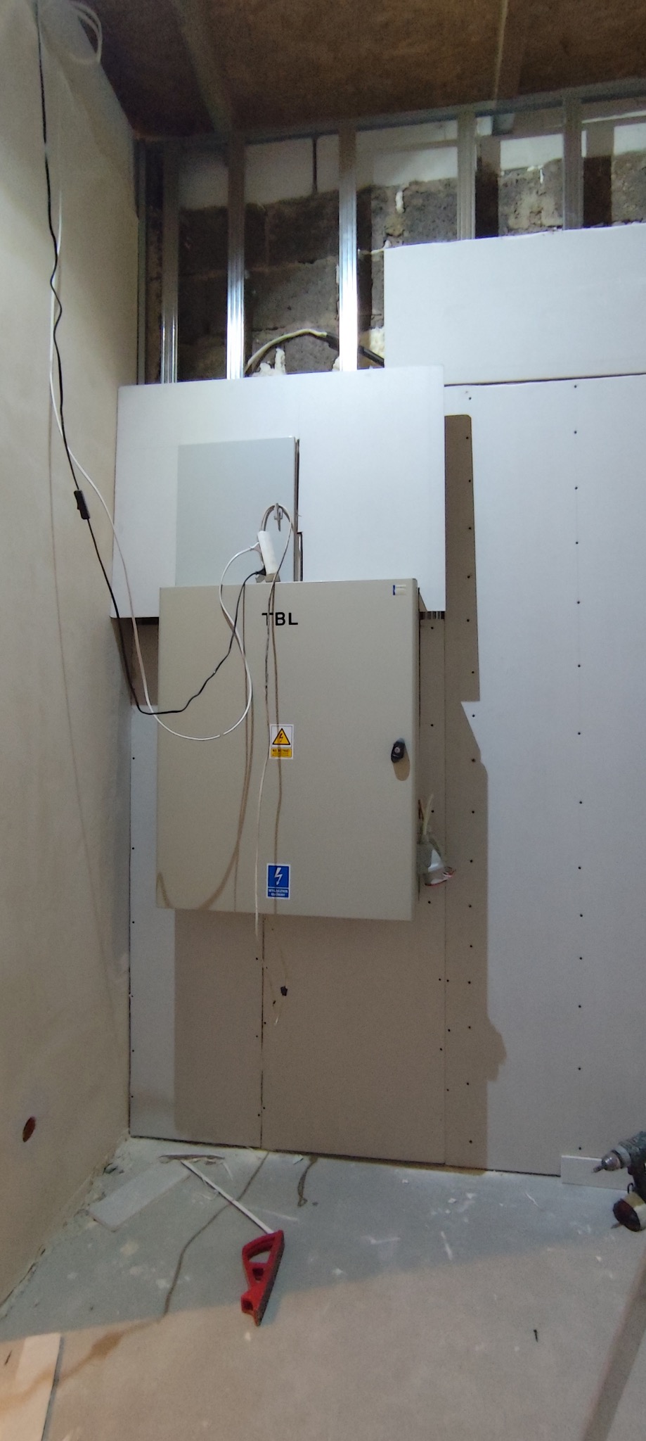 Montaż płyty karton gips na ścianie z widoczną skrzynką elektryczną, częściowo zakrytą płytami, metalowe profile konstrukcyjne w tle, na podłodze narzędzie do szpachlowania.
