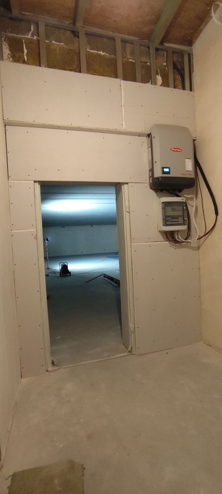 Montaż płyt kartonowo-gipsowych na ścianie z widocznym inwerterem Fronius i licznikiem energii, surowy stan wykończenia wnętrza z otwartym przejściem do kolejnego pomieszczenia.