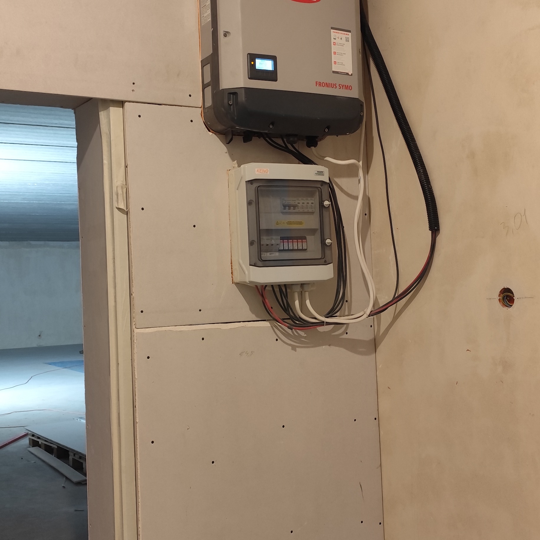 Montaż instalacji fotowoltaicznej: falownik Fronius Symo i skrzynka z bezpiecznikami na ścianie z płyt karton-gips, widoczne przewody i otwór w ścianie.