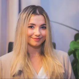 Portret uśmiechniętej blondynki w beżowej marynarce, z delikatnym naszyjnikiem z perłą, na neutralnym tle z rośliną doniczkową.