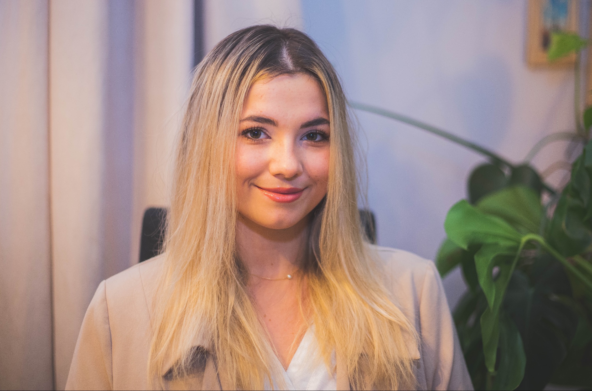 Portret uśmiechniętej blondynki w beżowej marynarce, z delikatnym naszyjnikiem z perłą, na neutralnym tle z rośliną doniczkową.