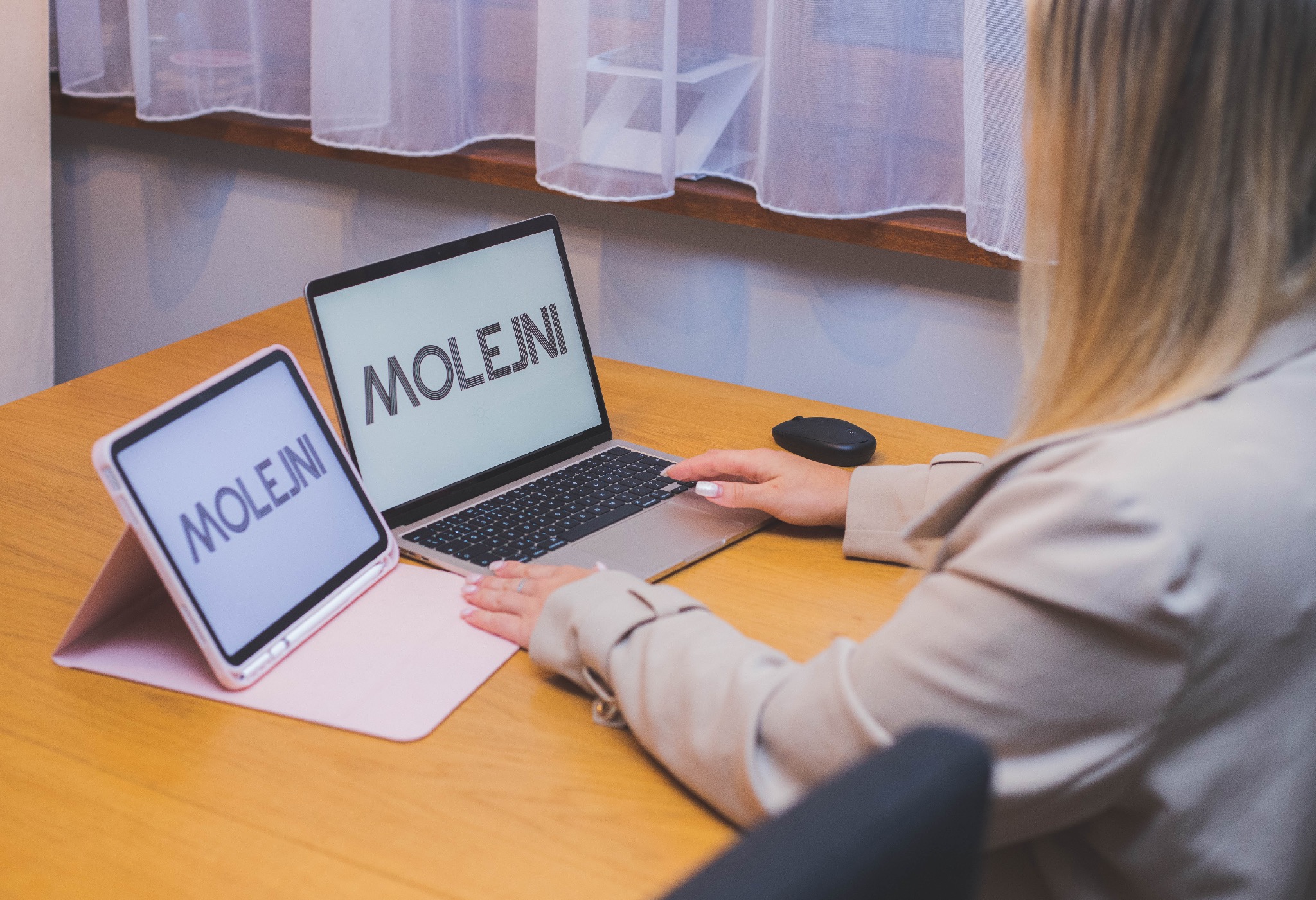 Kobieta w beżowym żakiecie pracuje na laptopie z wyświetlonym logo, obok leży tablet z tym samym logo na ekranie, widok zza pleców.