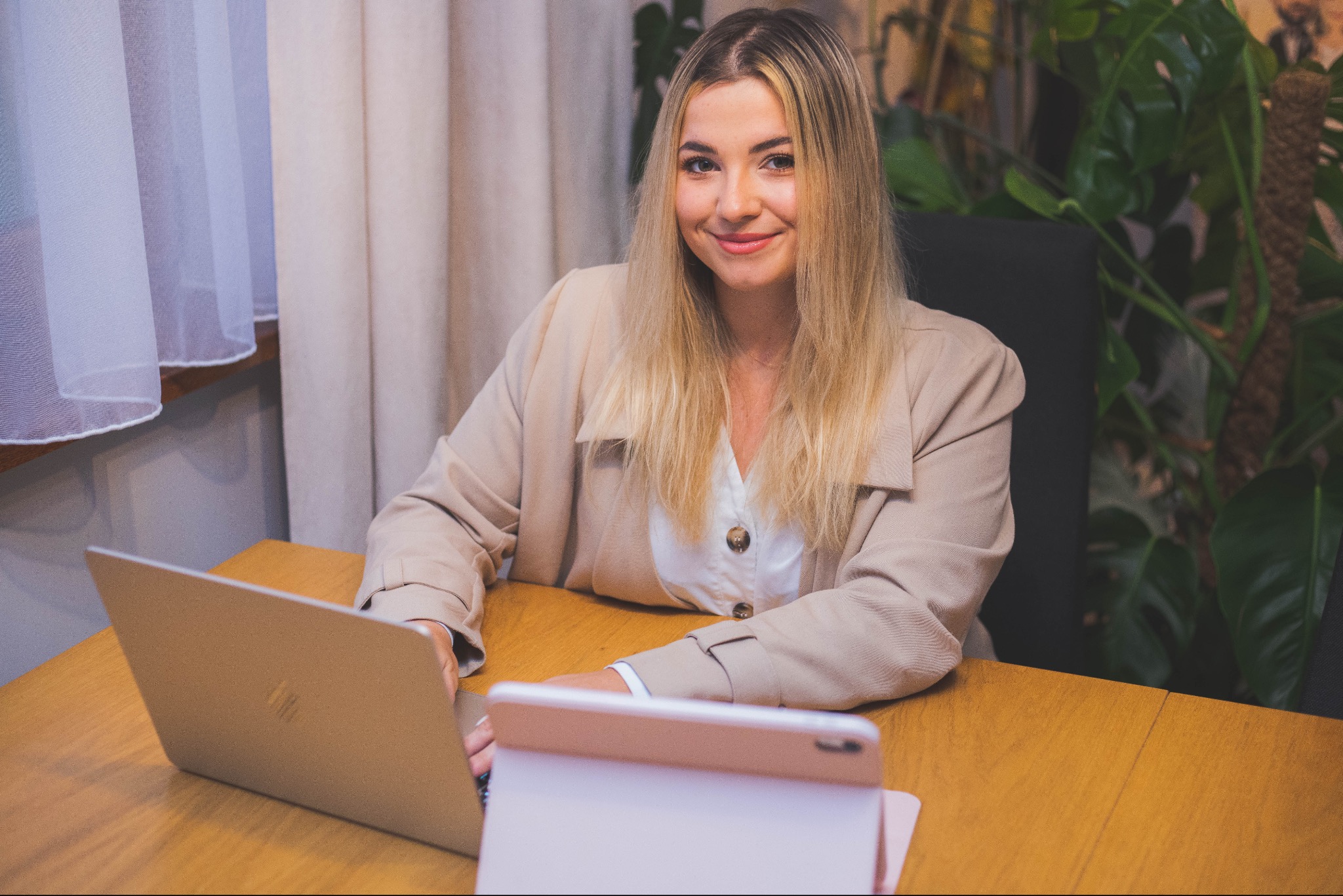 Uśmiechnięta blondynka w beżowej marynarce siedzi za biurkiem z laptopem i tabletem, w tle zasłony i roślina doniczkowa.