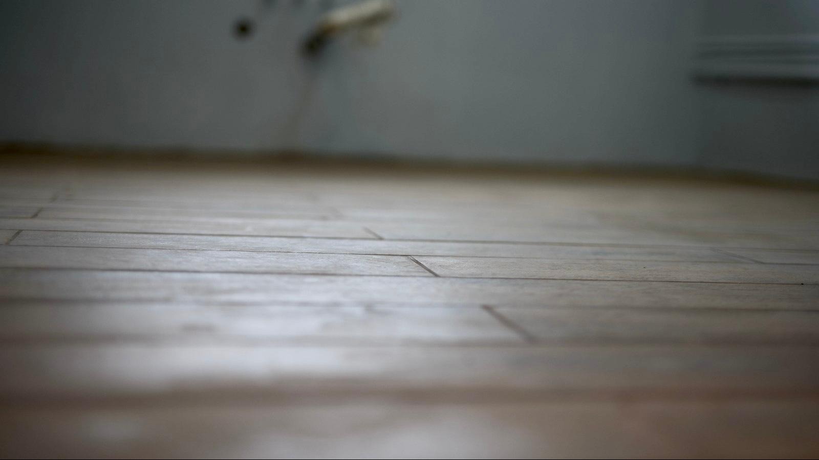 Ułożone panele podłogowe o jasnym, drewnopodobnym wzorze, wypełniające kadr od dołu do linii ściany w tle, z delikatnym efektem głębi ostrości.