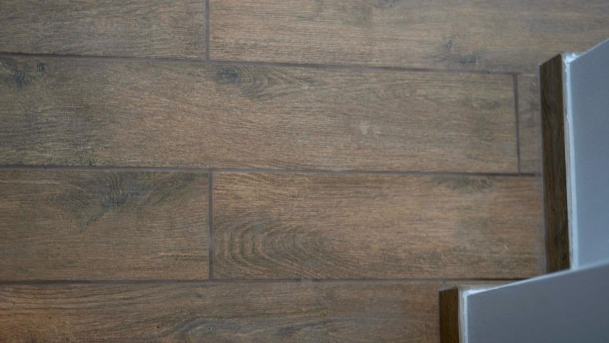 Drewnopodobne płytki ułożone poziomo na ścianie, z widoczną fugą i narożnikiem ściany. Ciepłe odcienie brązu i szarości.