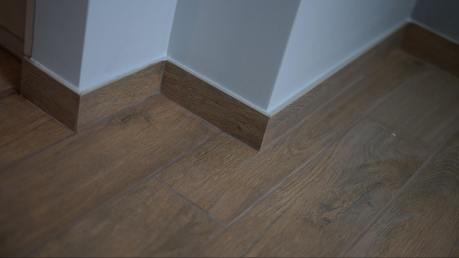 Ukończone brązowe panele podłogowe imitujące drewno, z dopasowanymi listwami przypodłogowymi przy jasnej ścianie. Detal wykończenia w rogu pomieszczenia.