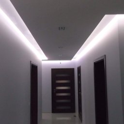 Promax - Korytarz z podwieszonym sufitem z oświetleniem LED, widoczne trzy ciemne drzwi z poziomymi przeszkleniami i punktowe oświetlenie sufitowe.