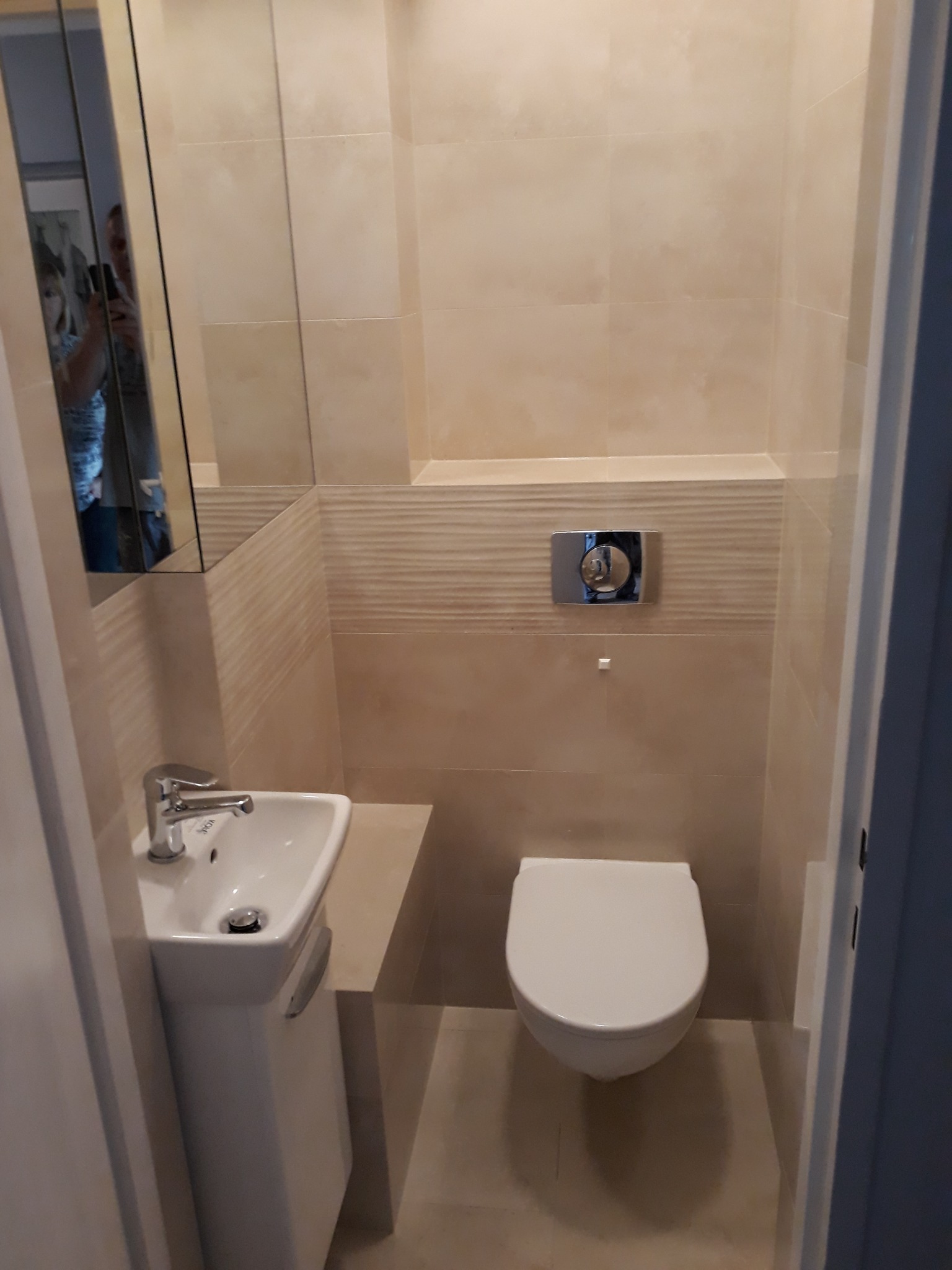 Mała toaleta dla gości z umywalką na postumencie, podwieszaną miską WC i beżowymi płytkami na ścianach, odbicie osoby robiącej zdjęcie w lustrze.