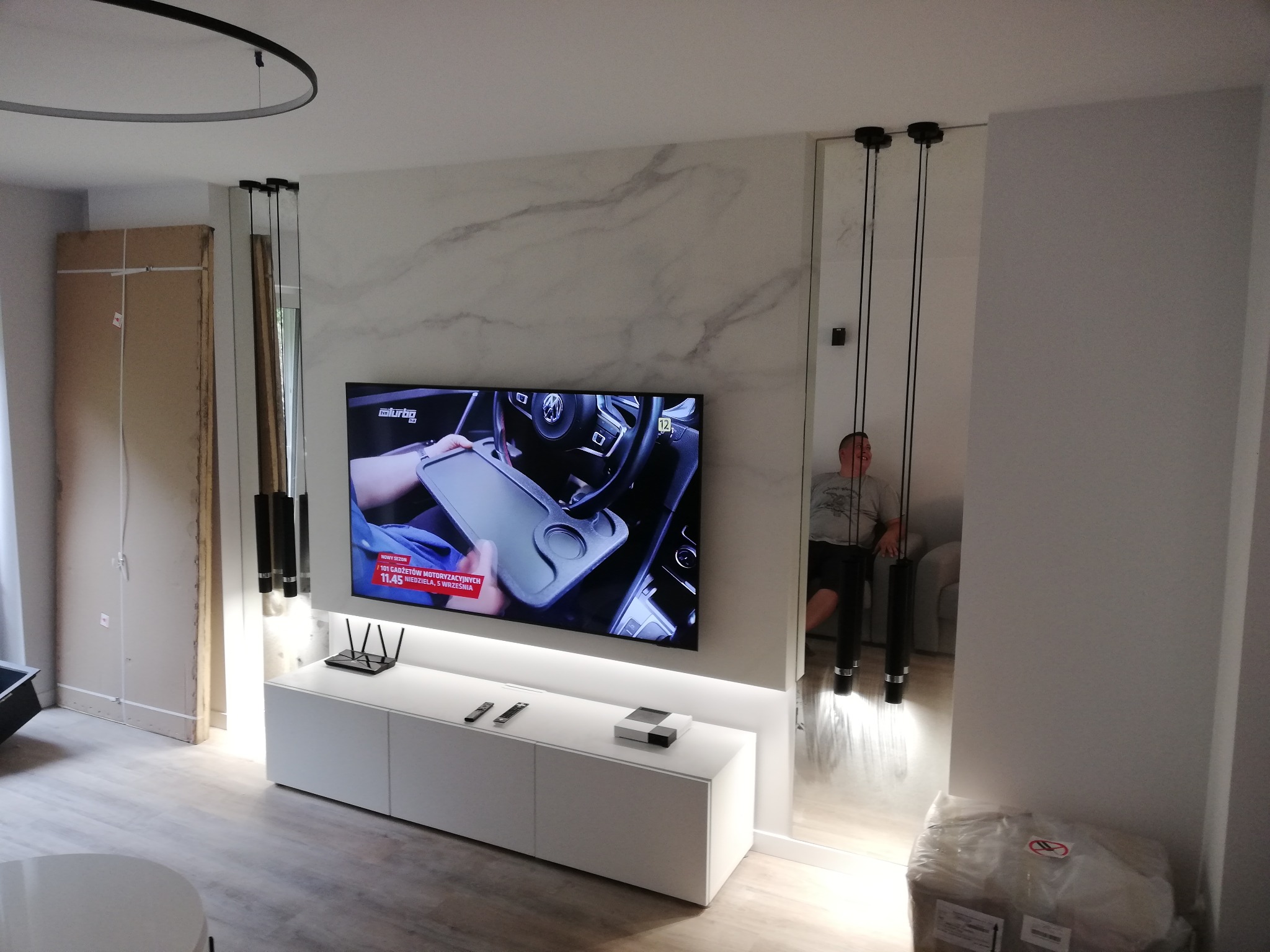 Telewizor zawieszony na ścianie imitującej marmur nad białą szafką RTV, z widocznymi elementami oświetlenia i fragmentem niezabudowanej ściany w tle, sugerujący remont.