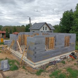 REKKER - Budowa domu w technologii szalunku traconego: ściany parteru wzniesione z szarych bloczków z widocznymi elementami deskowania i stemplami. W tle widoczny dom w stanie surowym zamkniętym.