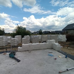 REKKER - Budowa domu z keramzytu: fundament z częściowo wzniesionymi ścianami, palety bloczków w folii w tle, narzędzia budowlane widoczne na płycie fundamentowej.