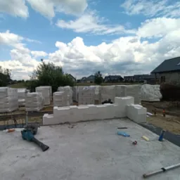 Budowa domu z keramzytu: fundament z częściowo wzniesionymi ścianami, palety bloczk&oacute;w w folii w tle, narzędzia budowlane widoczne na płycie fundamentowej.