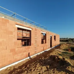 Surowy stan budynku z czerwonej cegły z widocznymi otworami okiennymi i drzwiami, zabezpieczony rusztowaniem na g&oacute;rze, otoczony piaskiem i ziemią, w tle zabudowania.