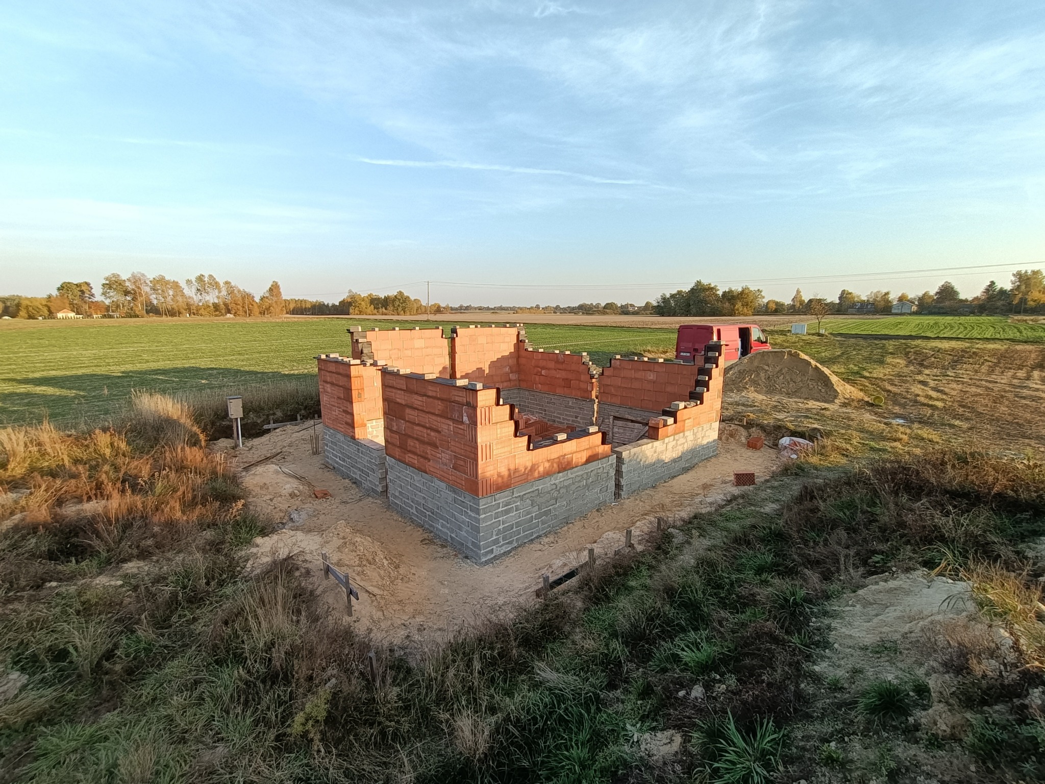 Wznoszenie ścian parteru domu z czerwonej cegły na fundamencie z szarych bloczków betonowych, widok z góry na plac budowy z polami uprawnymi w tle pod błękitnym niebem.