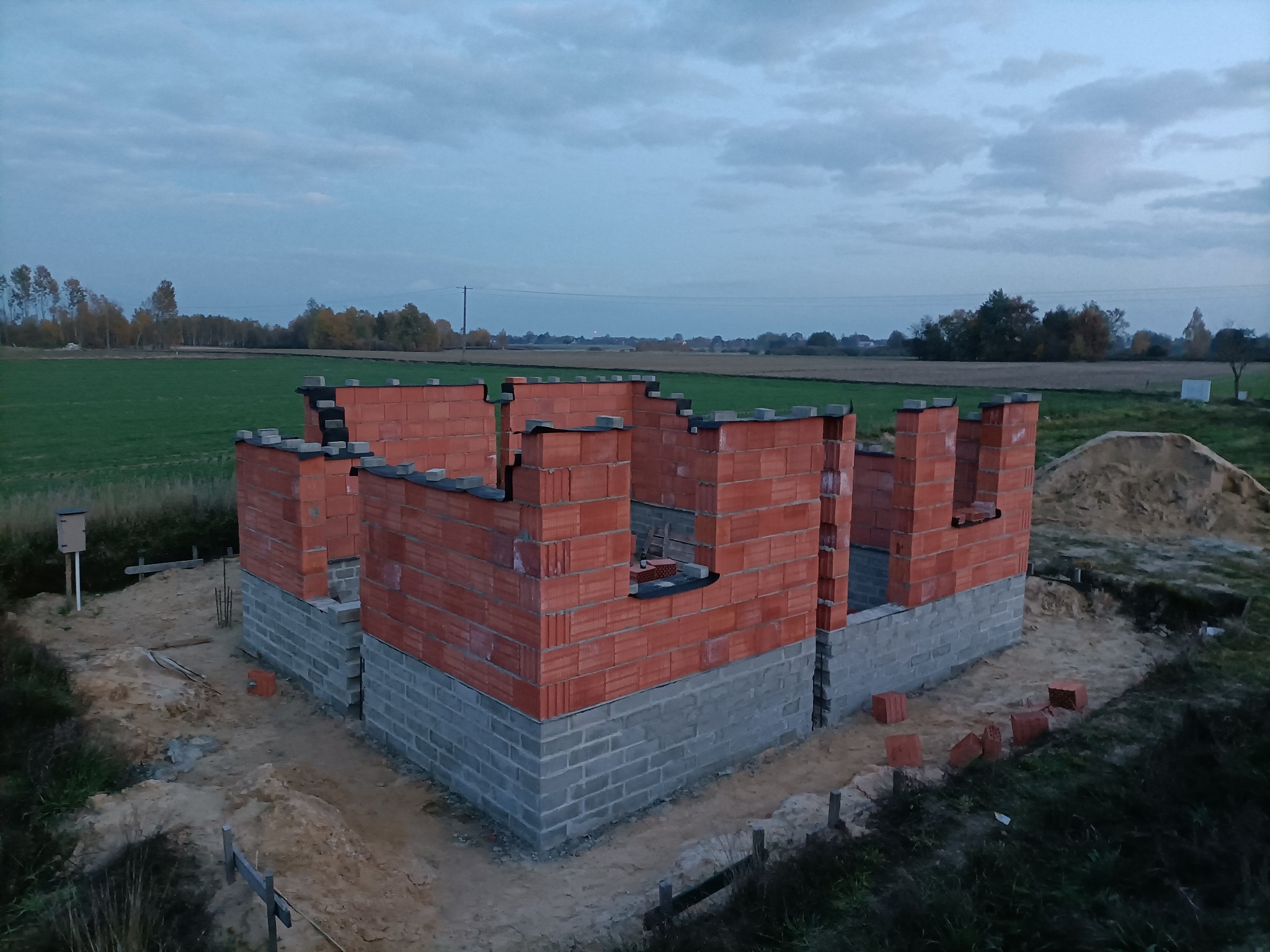 Wznoszone ściany parterowego domu murowanego z czerwonej cegły ceramicznej, widoczne szare bloczki fundamentowe i krajobraz pól w tle, pochmurne niebo.