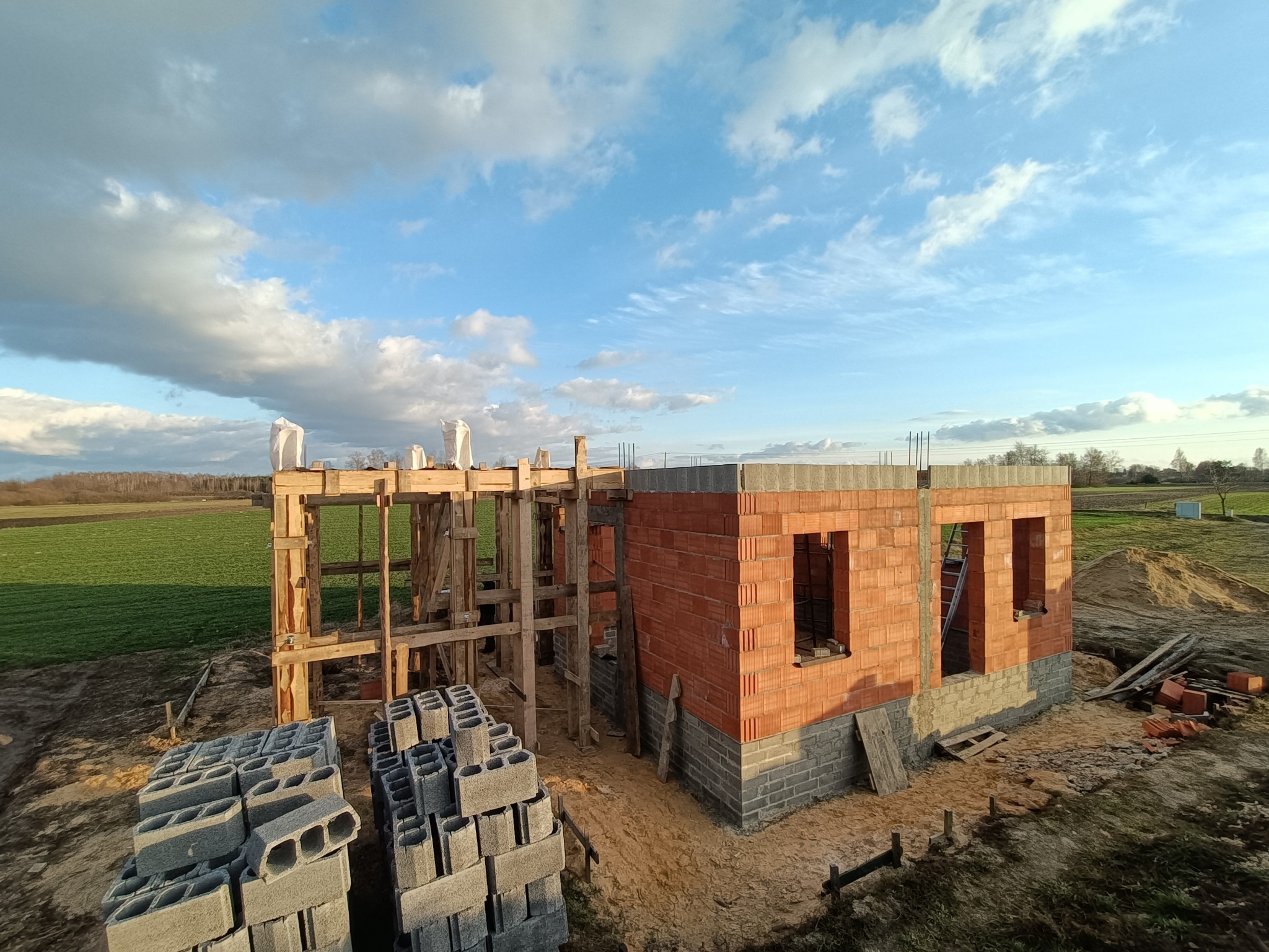 Wznoszenie ścian domu z czerwonej cegły i szarego bloczka, widoczny szkielet drewniany konstrukcji dachu, ułożone bloczki betonowe, tło stanowi zielone pole i błękitne niebo z chmurami.